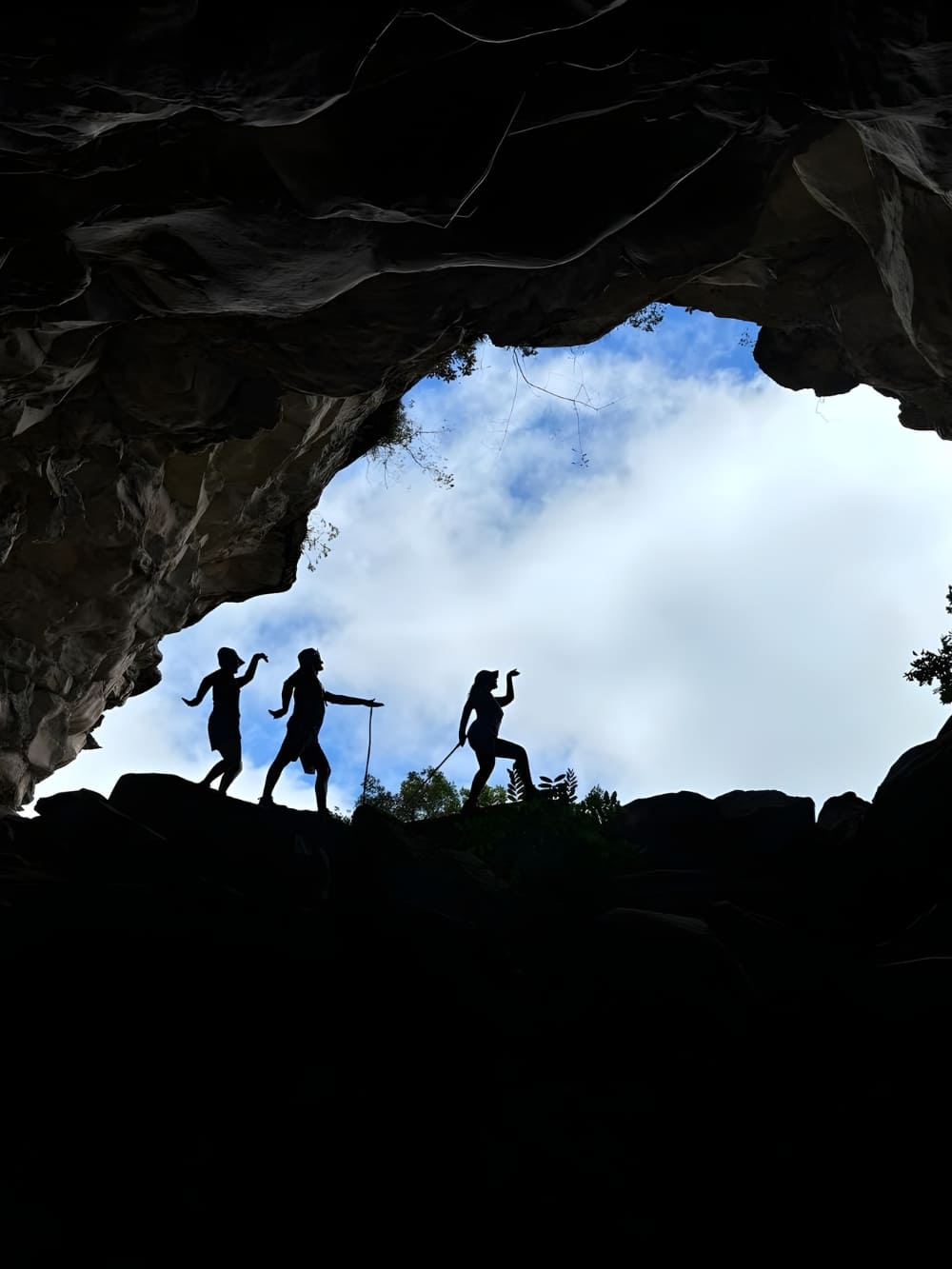 Silhouettes of three trekkers posing playfully at the entrance of a large dark cave in Chapada Diamantina, framed against a bright blue sky
