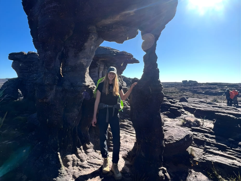 Mulher em pé sob uma enorme formação rochosa escura esculpida pela natureza, equilibrada sobre uma base estreita em uma paisagem de pedras sob sol forte. 