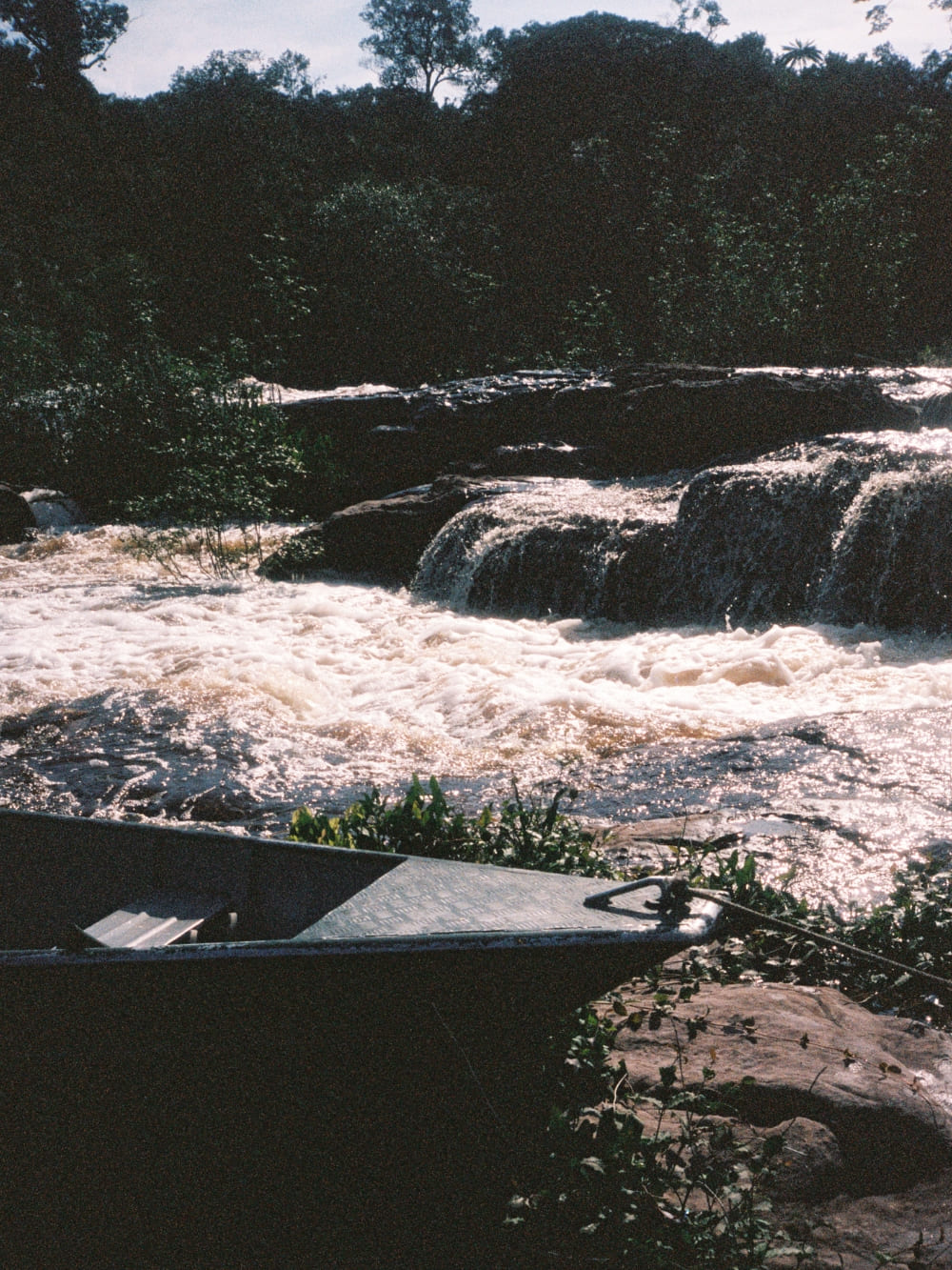 A small metal boat is moored at the rocky edge of an Amazonian river, with bright, fast-flowing water cascading over rocks in the background.