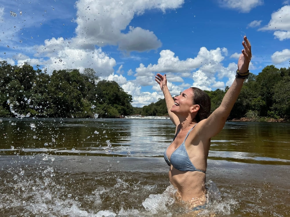 Lächelnde Frau badet im ruhigen Wasser eines von grünen Bäumen umgebenen Flusses und wirft mit ausgebreiteten Armen Wasser in die Luft.
