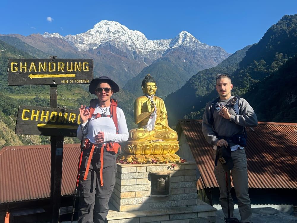 Frau und ein junger Mann mit Wanderrucksäcken lächeln neben einer goldenen Buddha-Statue, mit den majestätischen schneebedeckten Bergen des Himalaya im Hintergrund.