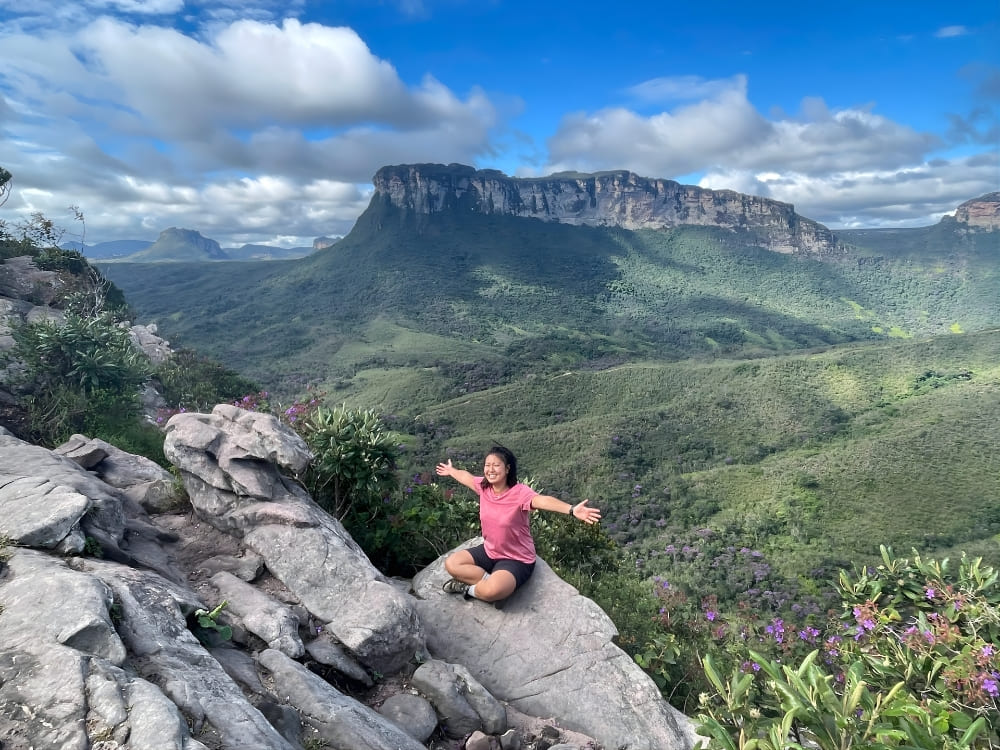  Mulher sorridente sentada de pernas cruzadas e braços abertos sobre uma rocha, admirando um vale verde com uma montanha imponente de topo plano ao fundo. 