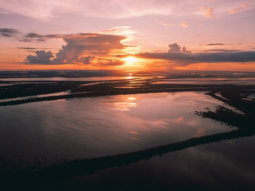 An aerial view captures a serene sunset over a flooded landscape, where strips of dark land separate wide pools of water reflecting the pink and orange sky.