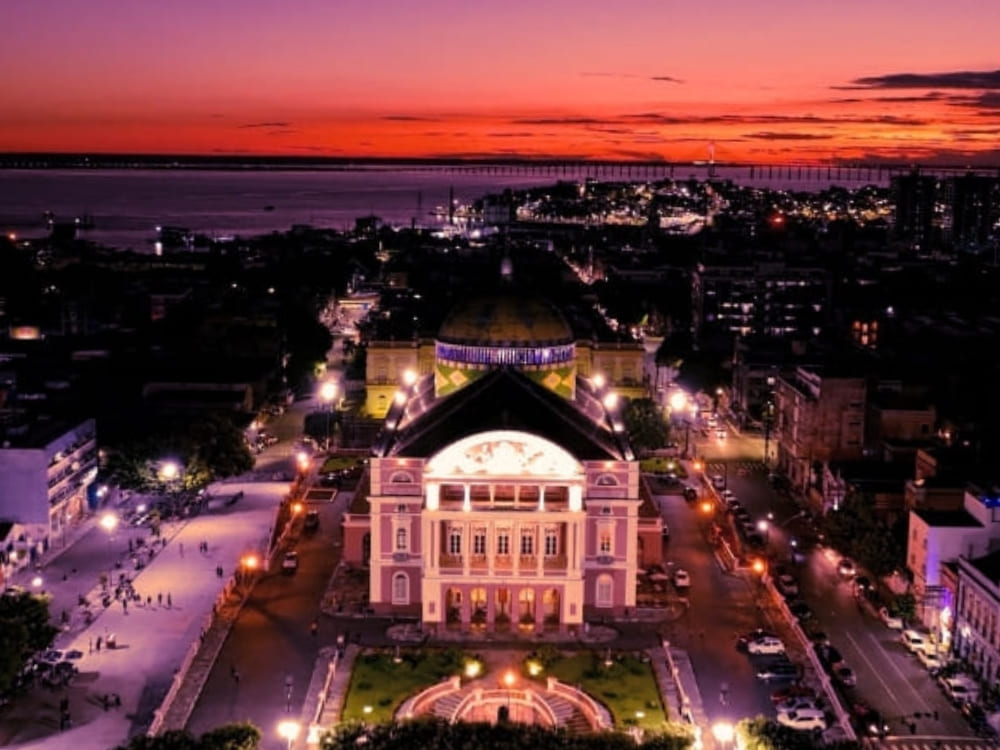 An illuminated historic theater with a colorful dome stands in a city center at dusk, with a river and bridge in the background.