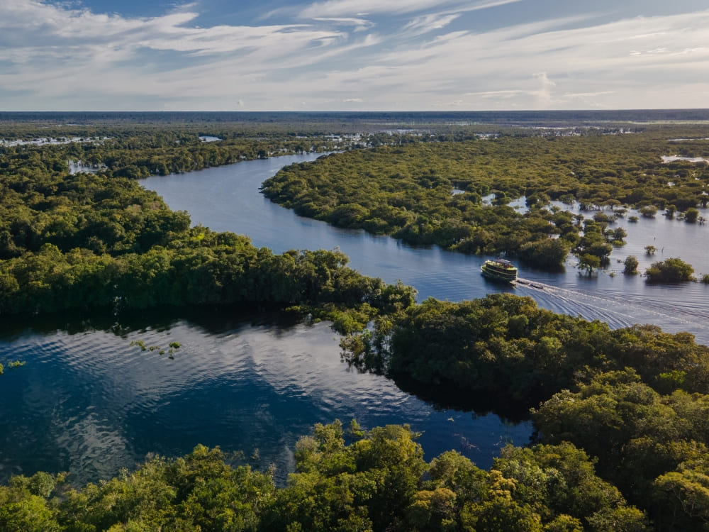 An aerial view of a river cruise boat navigating the dark waters of the Amazon river, surrounded by dense green rainforest under a partly cloudy sky.