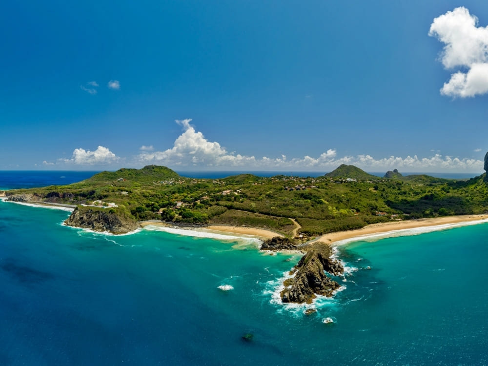 An aerial view shows a tropical island with lush green hills, sandy beaches, turquoise water, and a rocky peninsula.
