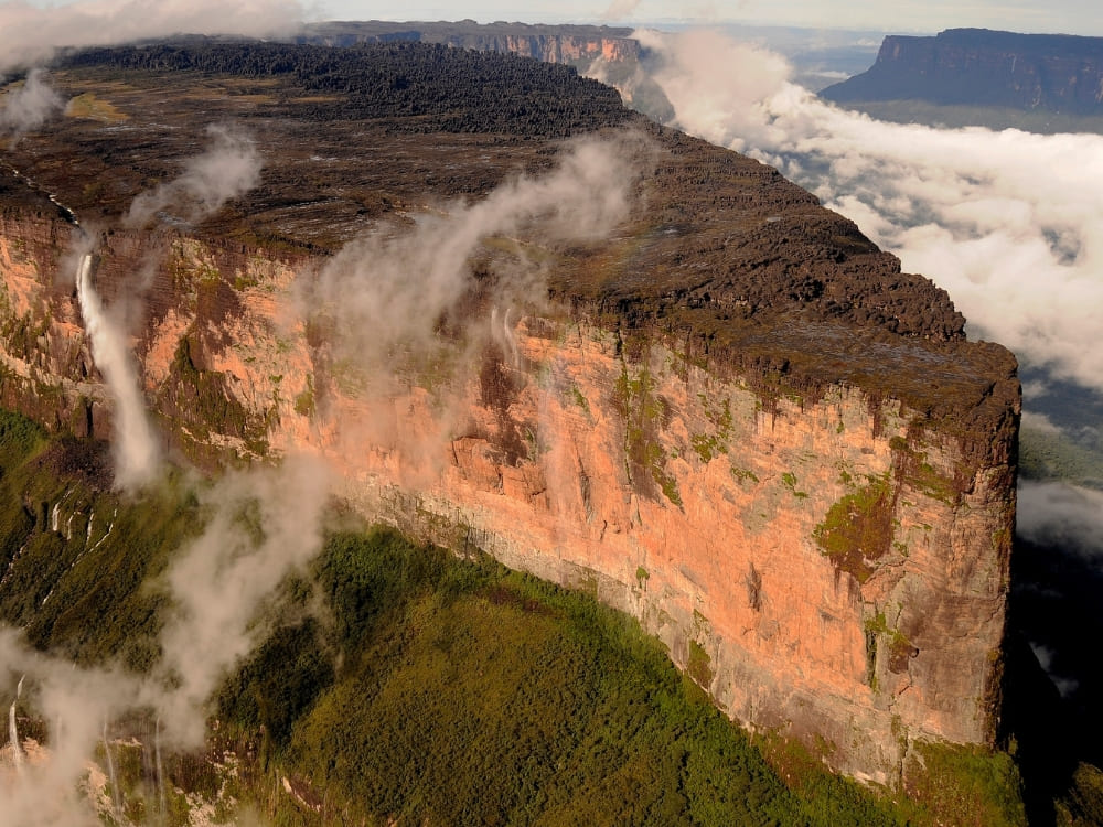 An aerial view shows a tall waterfall cascading down the sheer red cliff of a massive flat-topped mountain partially wrapped in clouds.