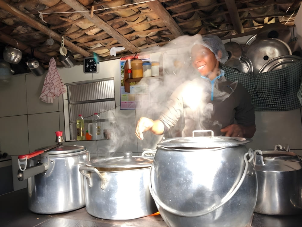  Host cooking with large pots in a rustic kitchen with steam, highlighting the legacy of native women who open the doors of their homes.