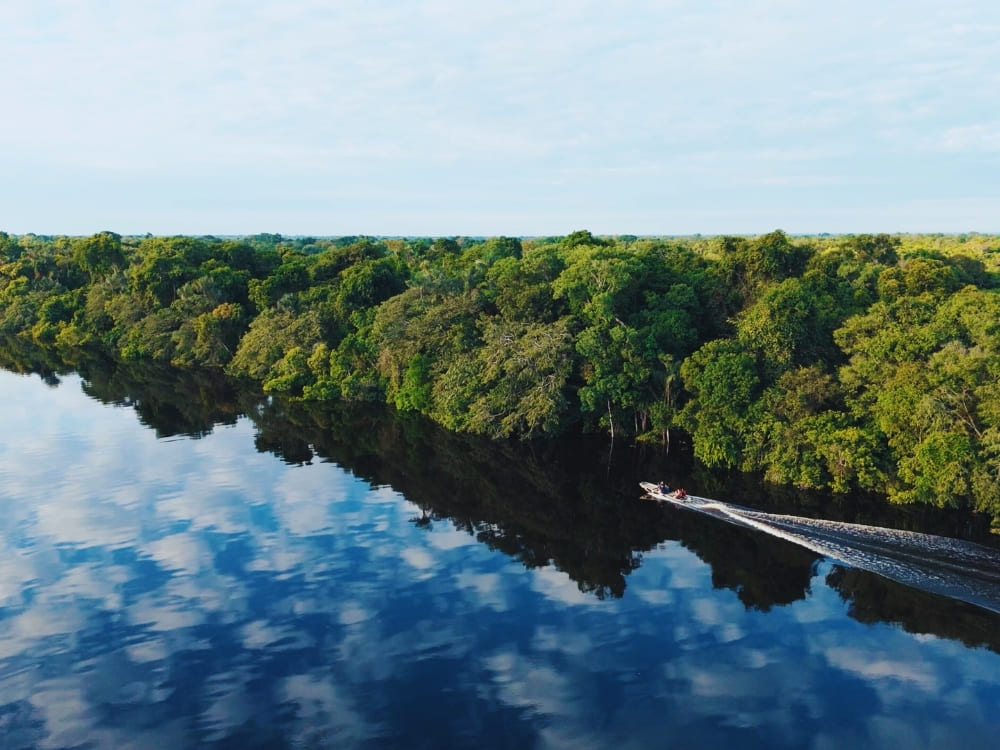 A small motorized boat travels down a river bordered by dense green jungle, leaving a wake on water that perfectly reflects the sky and clouds.