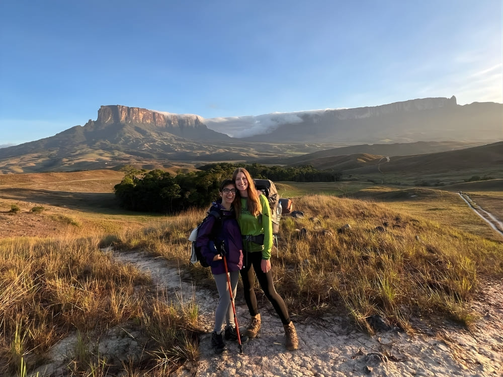 Two female hikers smile together on a grassy trail with the majestic Mount Roraima in the background. 