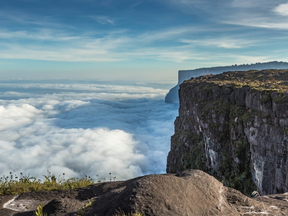 A towering flat-topped mountain cliff drops sharply into a thick, sweeping sea of white clouds under a bright blue sky.