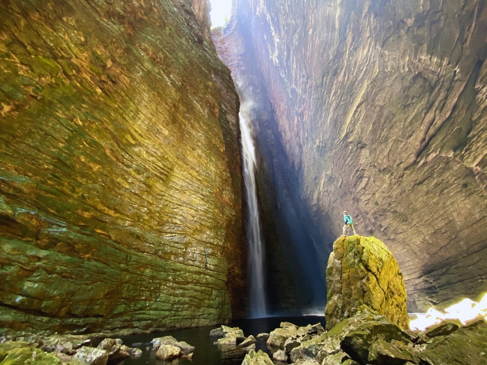 A person stands on a large mossy rock, looking up at a very tall, narrow waterfall cascading down a steep, brightly colored rock canyon.