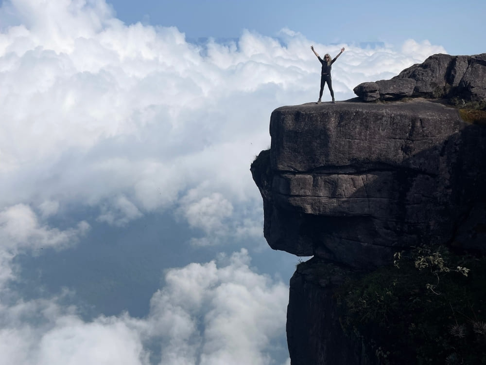 A hiker stands with arms raised in victory on a high, rocky cliff edge extending above a thick layer of white clouds.