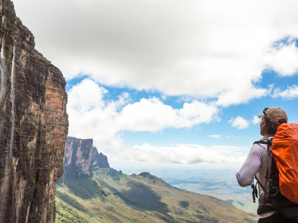 A hiker with an orange backpack looks out over a vast valley toward a distant, towering flat-topped mountain.
