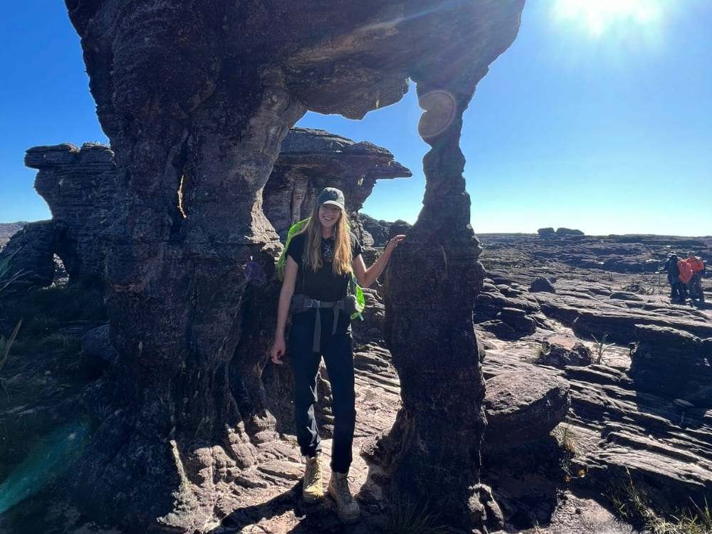 A female hiker with a backpack smiles while standing under a natural rock arch in a rugged, sunlit landscape.