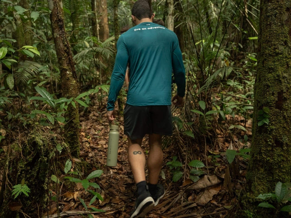A hiker carrying a water bottle walks along a dirt path through a dense, green tropical forest.