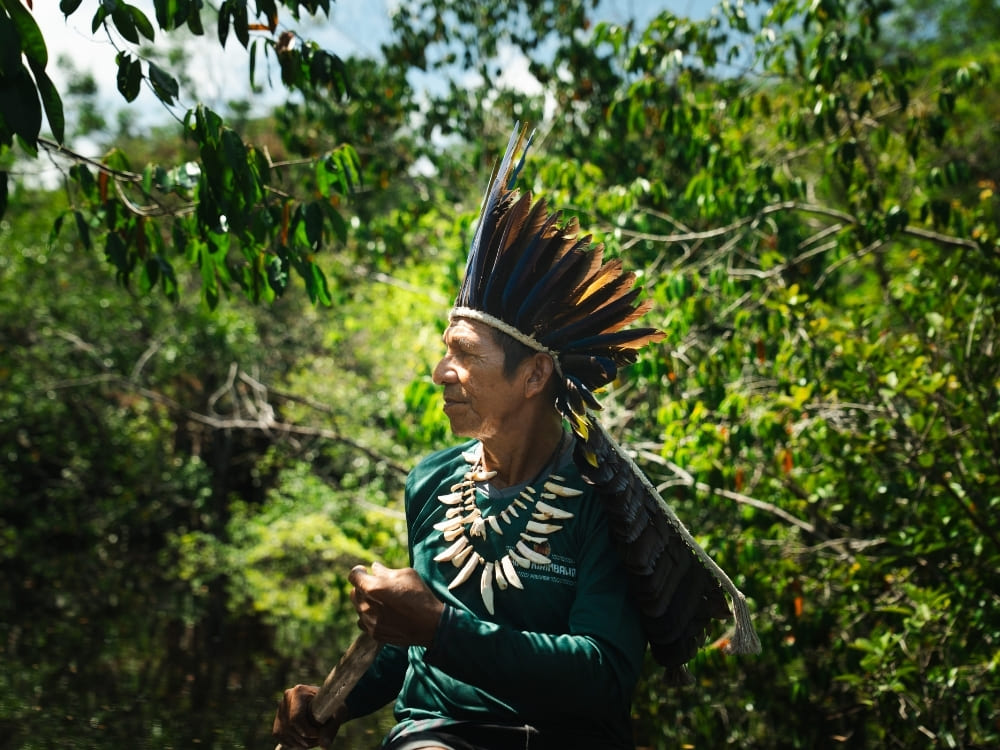 An indigenous man wearing a large feathered headdress and a tooth necklace paddles a wooden boat through a dense, sunlit green forest.