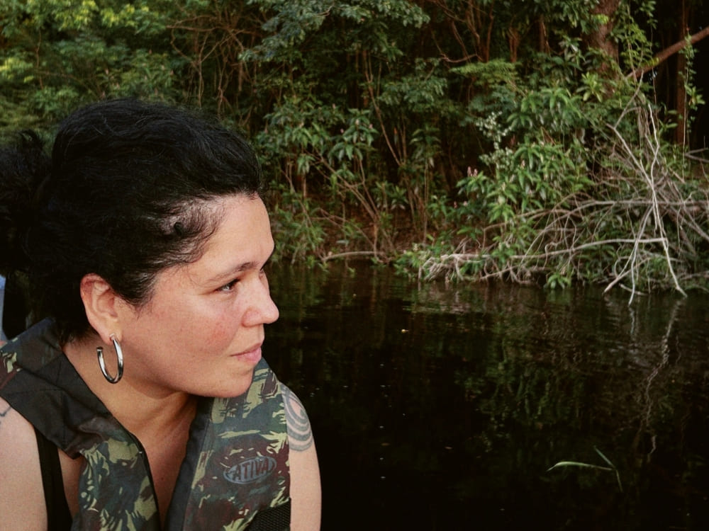 Eine dunkelhaarige Frau mit Creolen und einer getarnten Rettungsweste sitzt in einem kleinen Boot und blickt nachdenklich auf die üppige Ufervegetation.