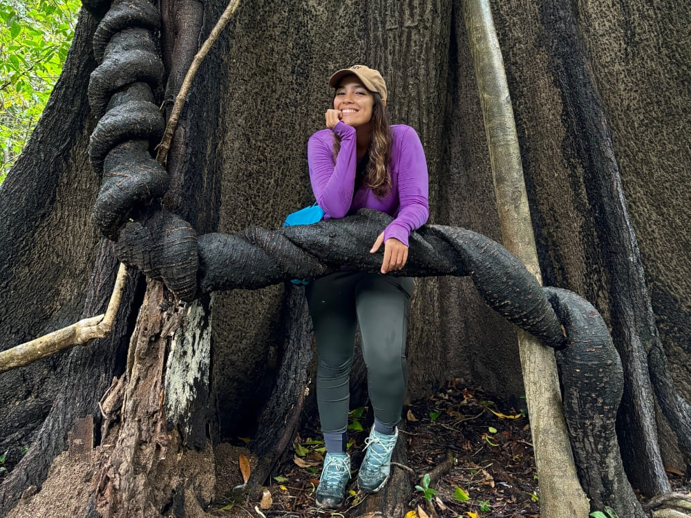 Mariana rests on a giant twisted tree root while exploring the dense greenery of the Amazon jungle. 