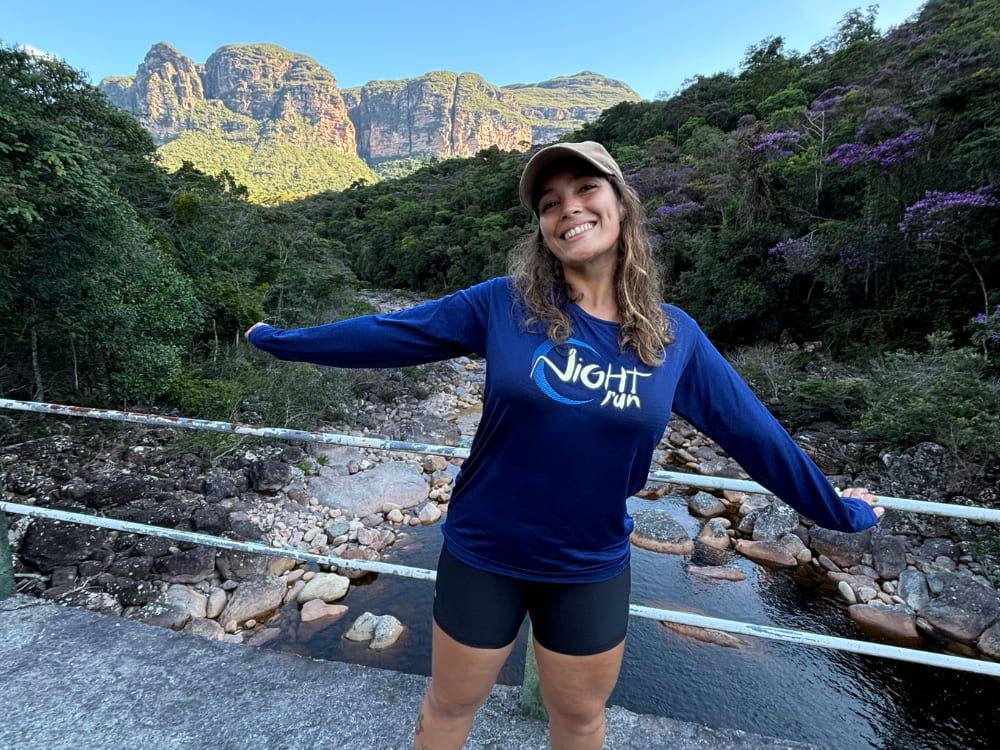 A smiling Mariana stands with arms outstretched on a bridge over a rocky river with the rugged mountains of Vale do Pati in the background.