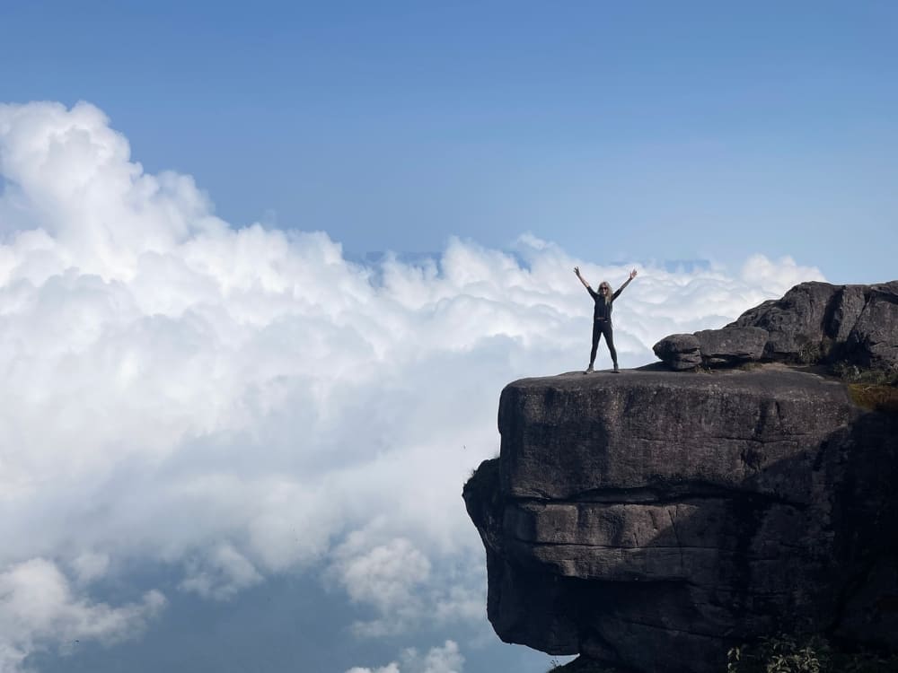 Silhouette of a woman with her arms raised in a sign of victory on the edge of the rocky cliff of Mount Roraima, above the white clouds.