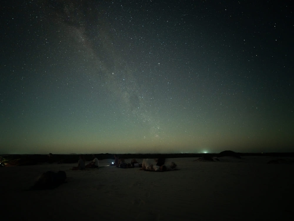 A group of people sit and relax on dark sand dunes at night under a clear sky filled with stars and the Milky Way.