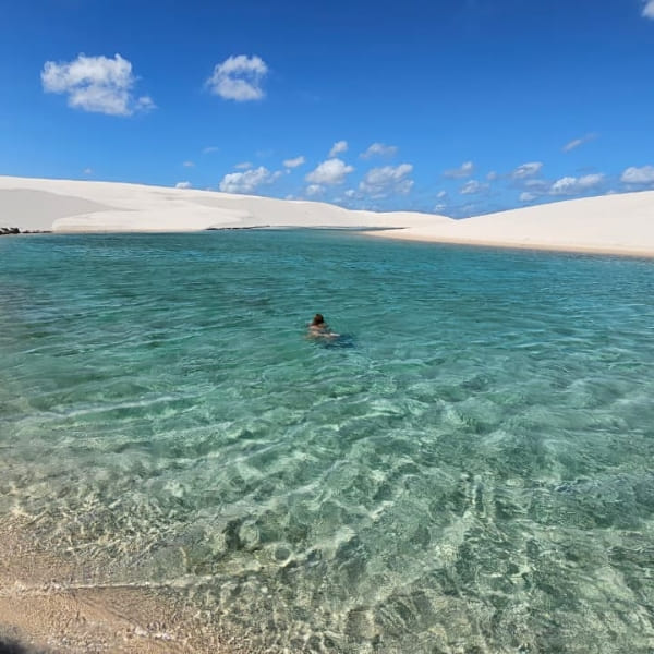 Eine Person schwimmt in einer weitläufigen Lagune mit transparentem, blauem Wasser zwischen den großen Sanddünen von Lençóis Maranhenses.
