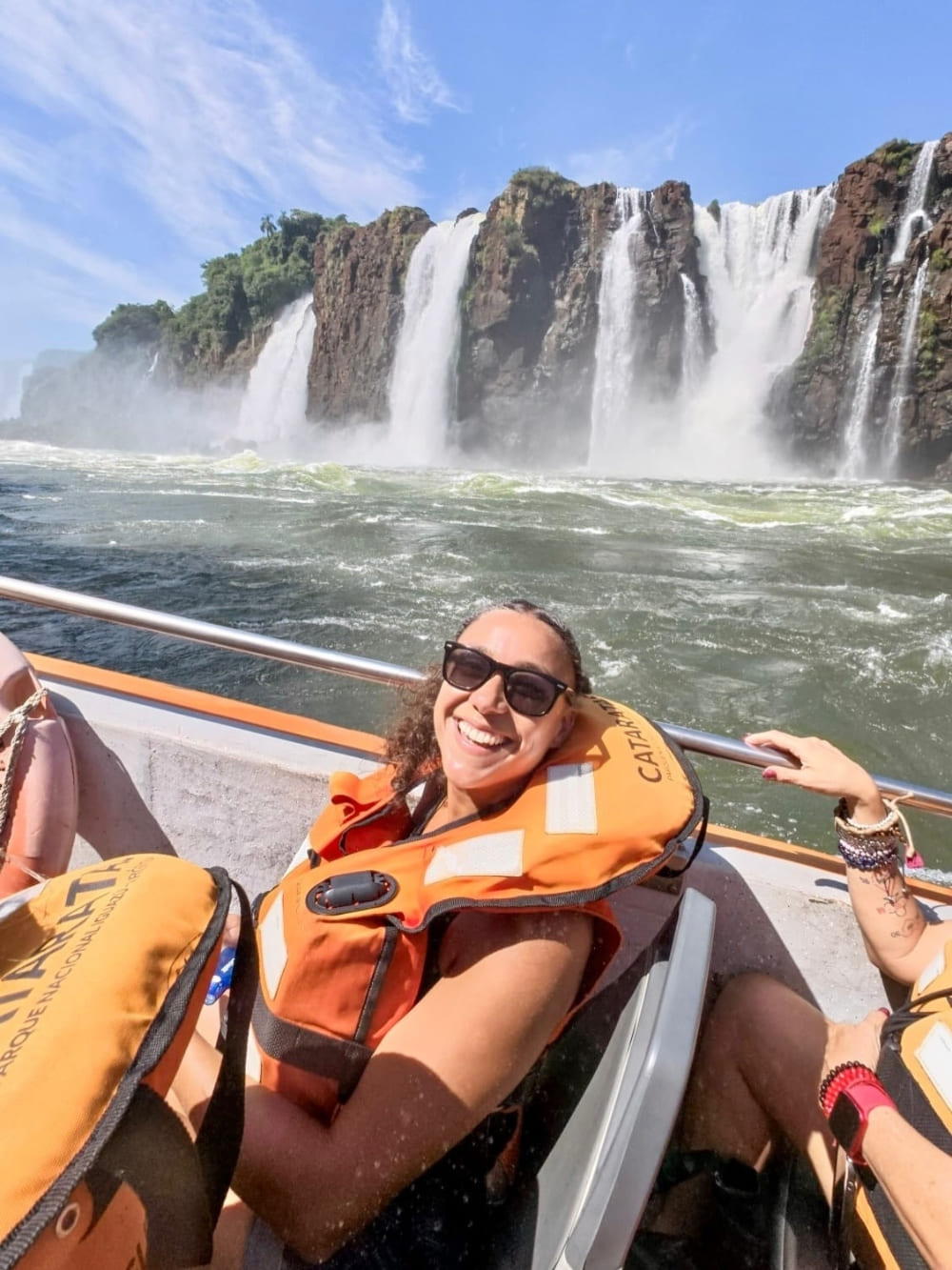 Samantha Sage wears sunglasses and an orange life jacket while smiling on an adventure boat ride near massive cascading waterfalls in Brazil.