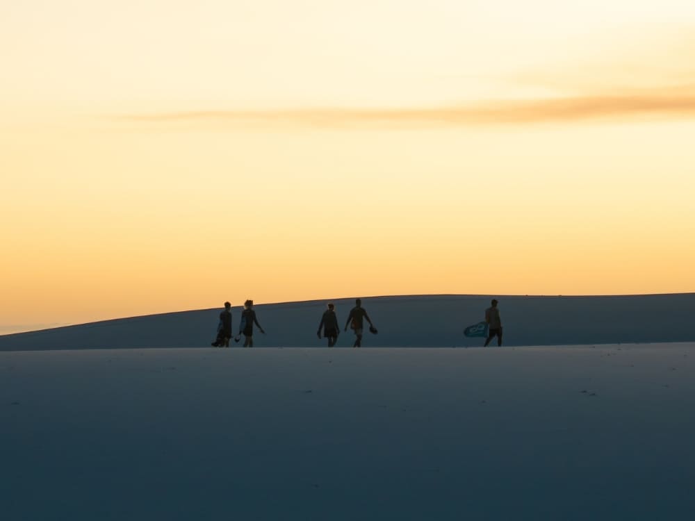 Silhouettes of five people walk across smooth, white sand dunes against a vibrant yellow and orange sunset sky.