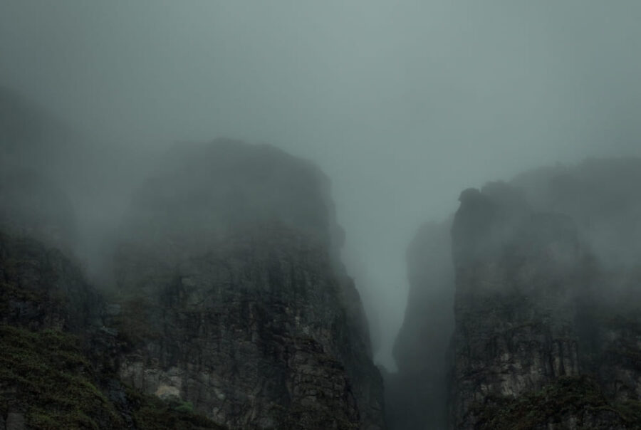 Steep, dark rocky mountain cliffs are partially obscured by thick, gray fog and low-hanging clouds.
