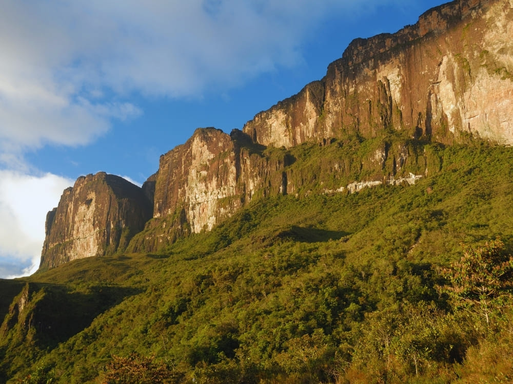 Sunlit flat-topped mountains covered in lush green vegetation rise against a blue sky.