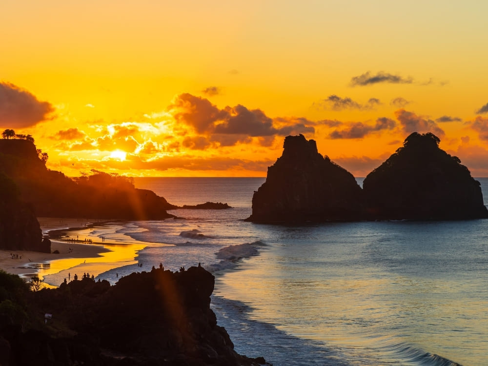A vibrant golden sunset illuminates a calm beach, silhouetting people and two massive, twin rock formations rising from the ocean.