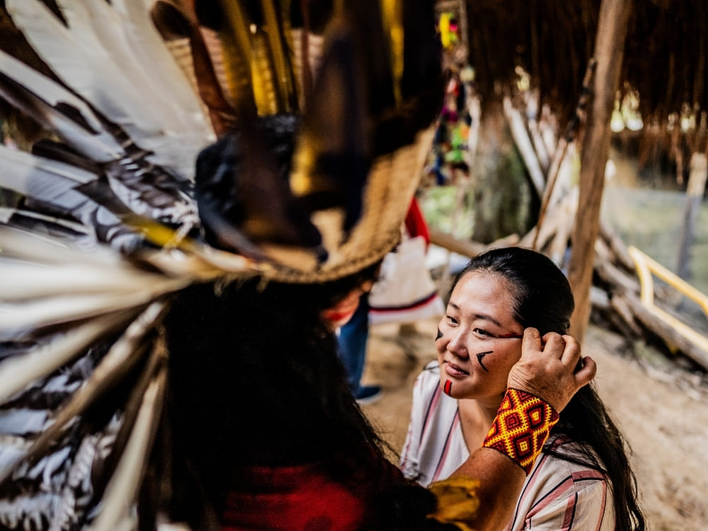 A smiling female tourist receives traditional red face paint from an indigenous person wearing a colorful feathered headdress.