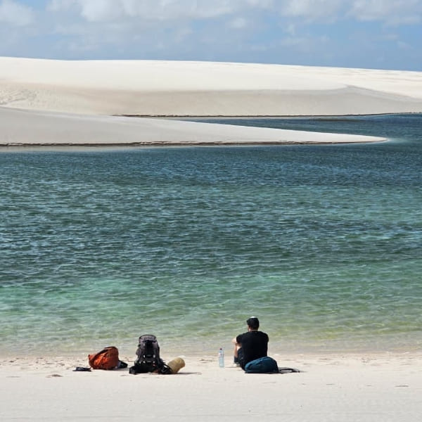 Ein Rucksacktourist ruht sich im weißen Sand aus und blickt auf das ruhige grüne Wasser einer Lagune in Lençóis Maranhenses.