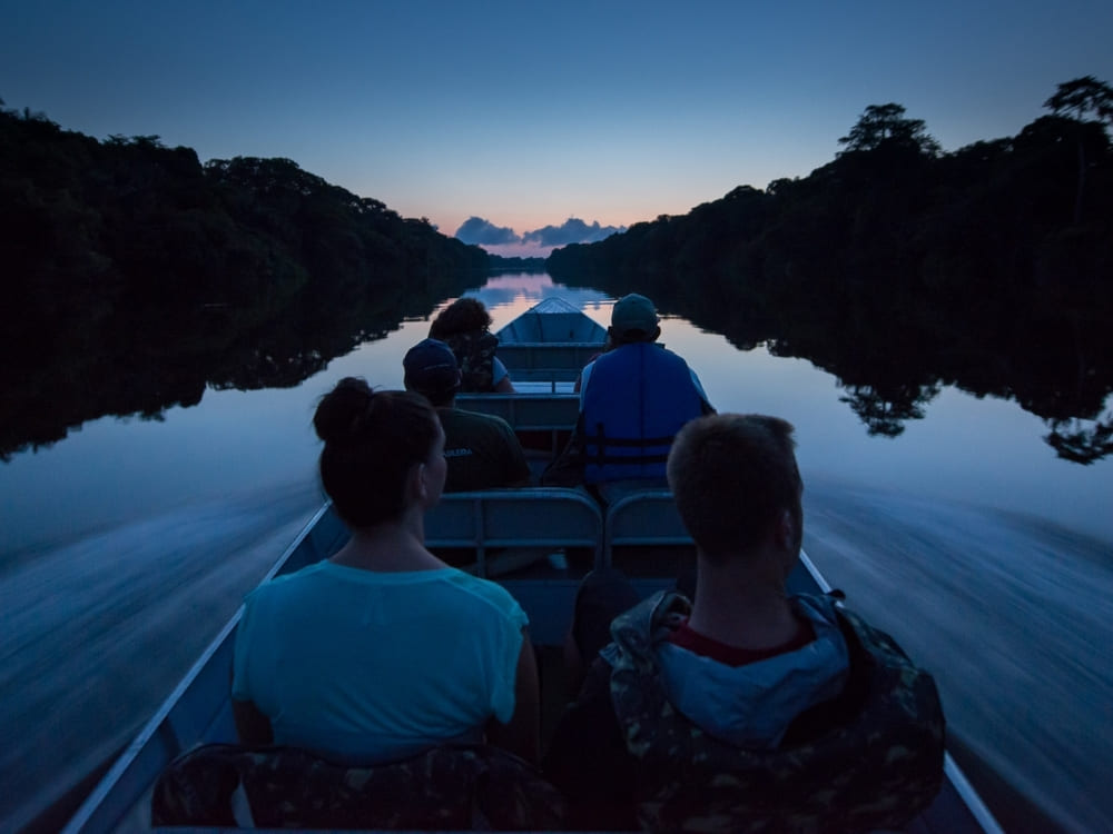 Passengers ride in a small boat down a calm, dark river bordered by silhouetted trees during twilight.