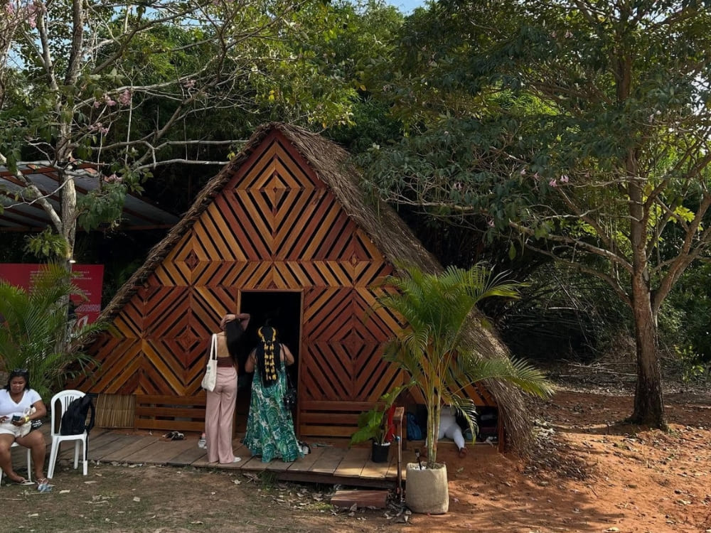 Two women enter a traditional triangular structure with a thatched roof and geometric wooden facade, surrounded by trees and reddish soil in the Amazon.