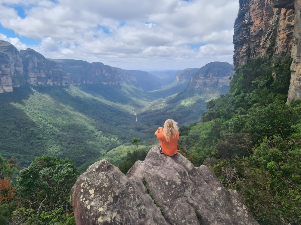 Woman in an orange jacket sitting on a high rock, admiring the panoramic view of a vast green valley in Chapada Diamantina.