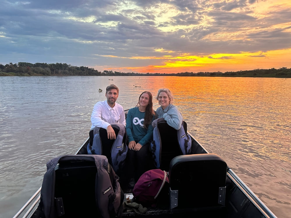 Three people smiling in a small boat navigating a river during a vibrant sunset, experiencing sustainable tourism tours.