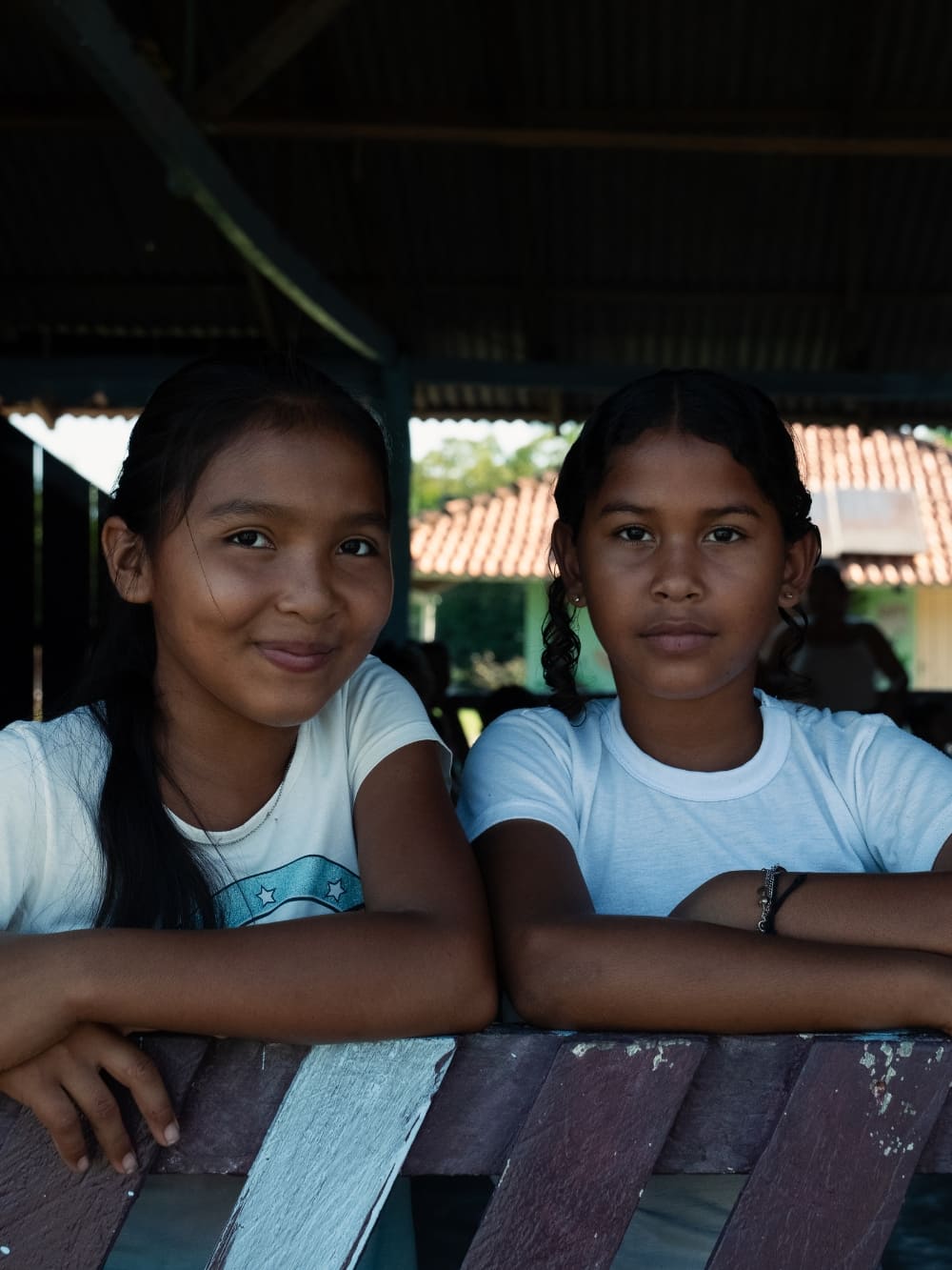 Two local student girls smile warmly while leaning on a wooden fence in an Amazon riverside village.