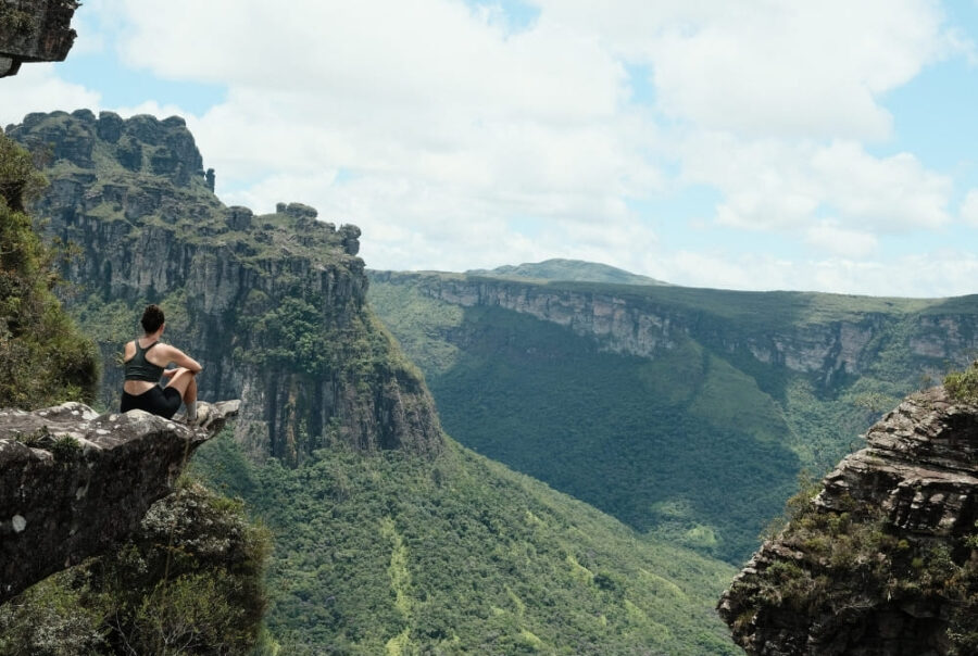 A woman sits on the edge of a steep rocky cliff, taking in the view of a massive, green canyon and distant mountains.