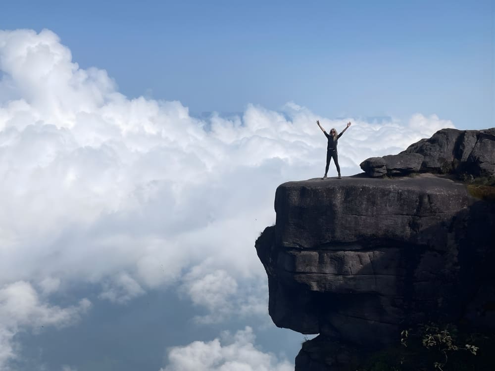  Solo female traveler standing with arms raised in victory on a high mountain cliff edge above the clouds in Brazil.