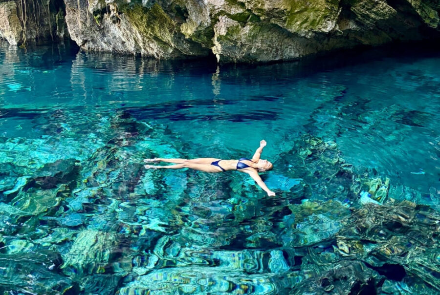 A woman in a dark swimsuit floats peacefully on her back in crystal-clear, bright blue water near a rocky cave wall.