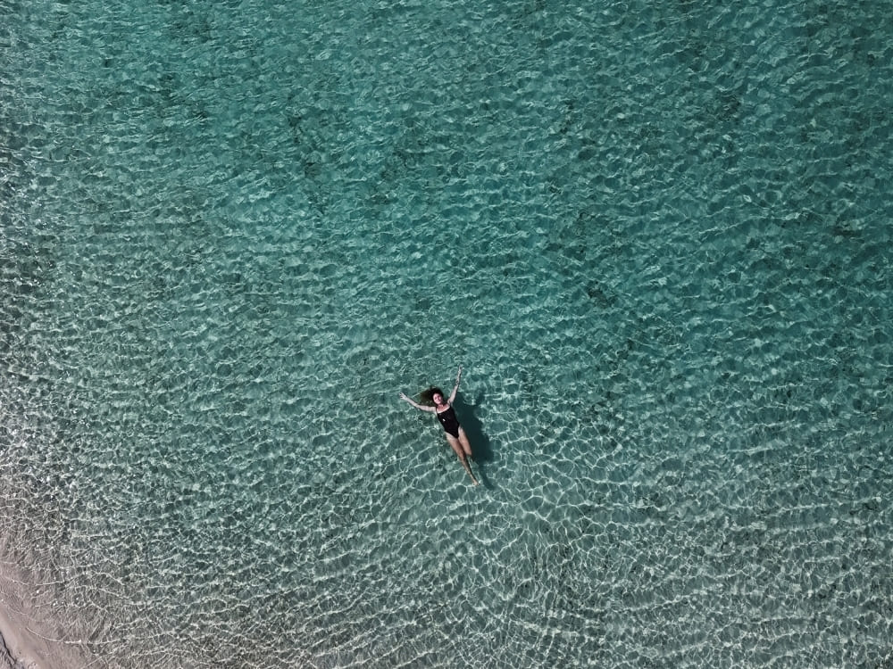An aerial view shows a woman in a dark swimsuit floating peacefully on her back in clear, bright turquoise water.