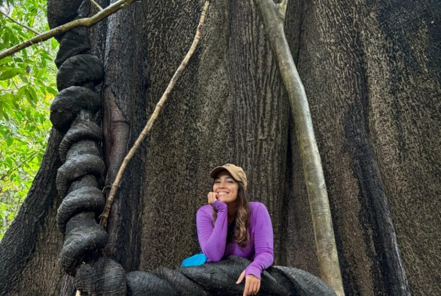 A smiling woman leans against thick, twisted vines at the base of a massive tree trunk in a tropical forest.