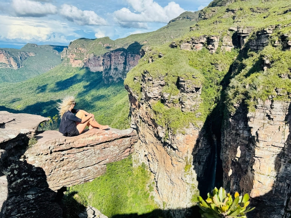 A blonde woman sits on a protruding rock ledge, looking out over a deep canyon and lush green valley under a sunny sky.
