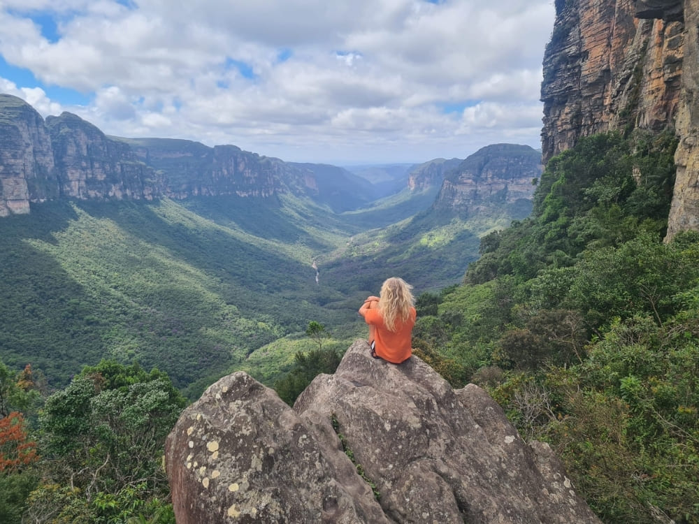 A woman sits on a rocky outcropping, looking out over a vast, lush green canyon bordered by steep vertical cliffs.