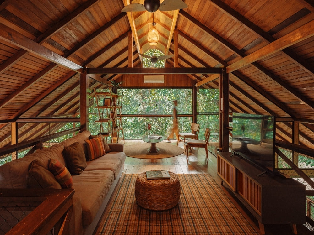 The interior of a rustic wooden eco-lodge features a living room and a glass wall looking out into a dense green jungle.