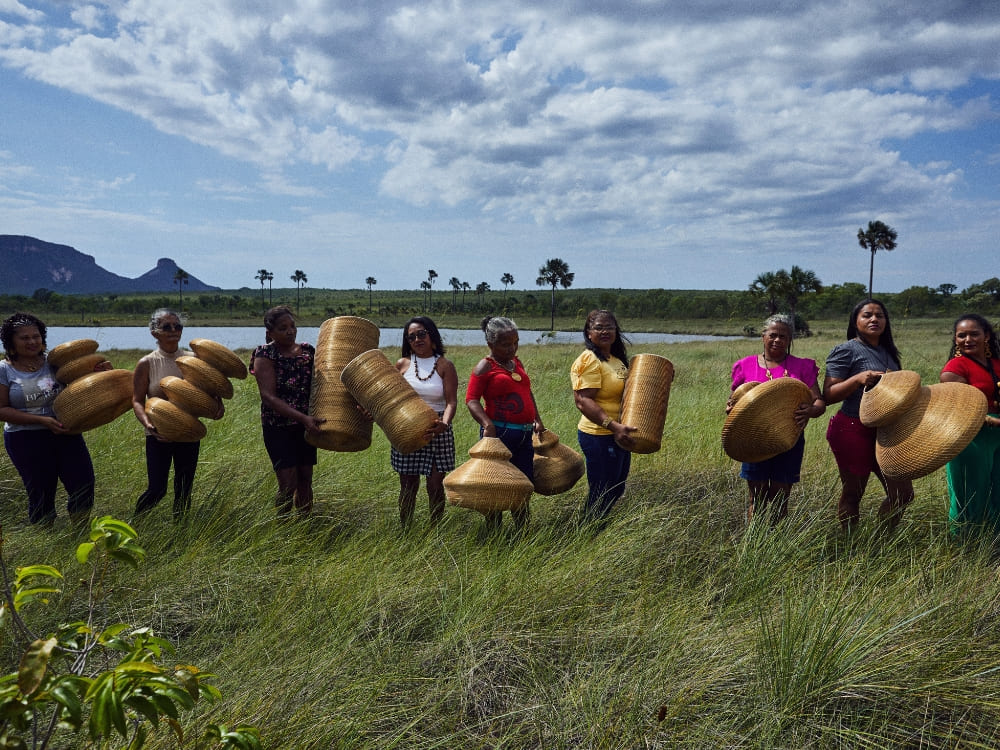 Um grupo de mulheres de pé em um campo verde exuberante sob um céu nublado, segurando artesanatos tecidos feitos de Capim Dourado na região do Jalapão.