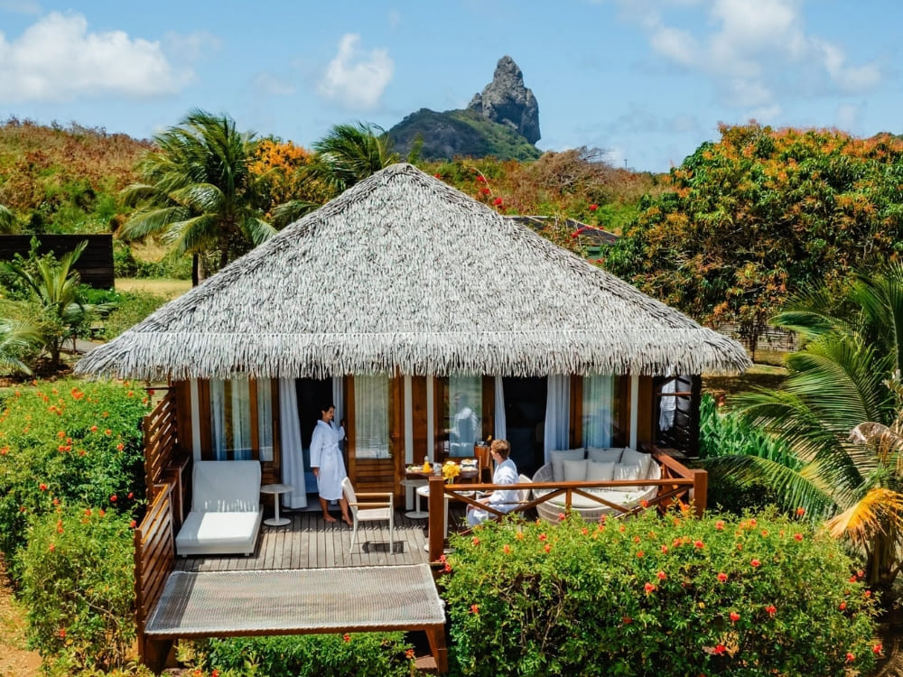 Couple in white bathrobes relaxing on the private deck of a rustic thatched-roof bungalow with views of Morro do Pico at a top hotel in Fernando de Noronha.