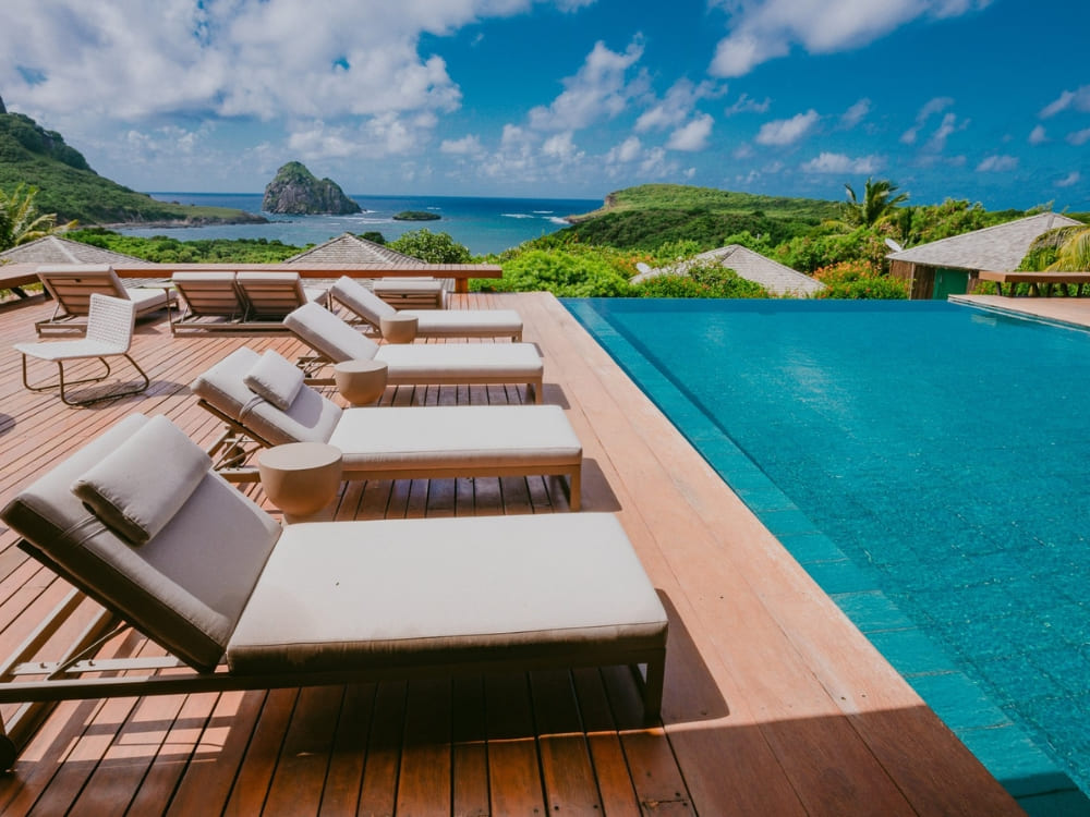  Wooden deck with comfortable lounge chairs beside an infinity pool overlooking the ocean and Morro do Pico at a hotel in Fernando de Noronha